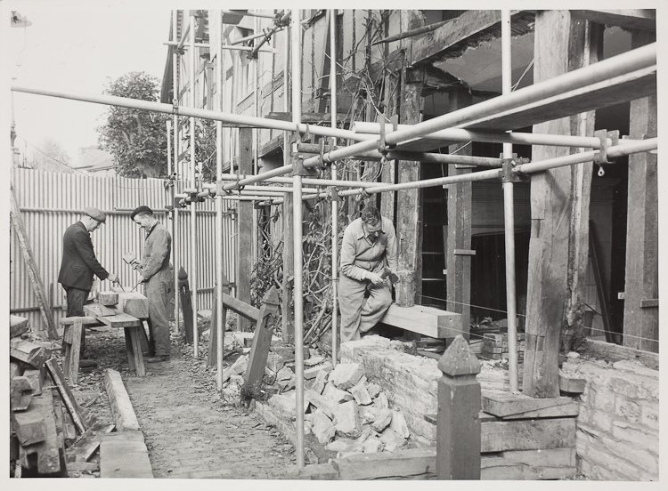 Behind a man working at beams from which the panels have gone a tangle of wattle hangs down. Scaffolding is across the house, there are piles of bricks and rubble, and in the background two men are examining a beam on a workbench.