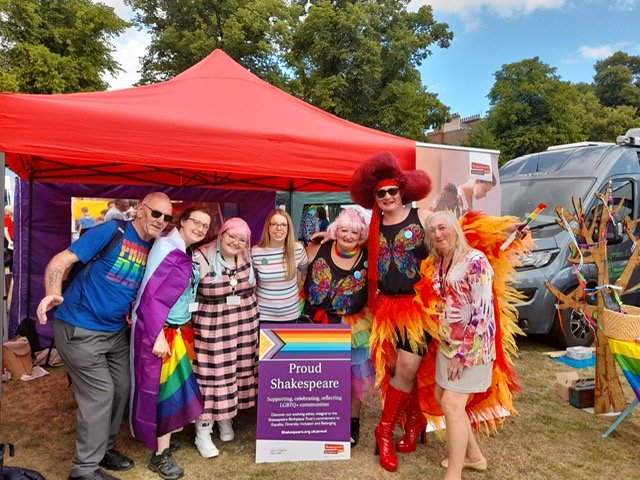 Shakespeare Birthplace Trust staff and volunteers pictured with visitors to the SBT stand at Warwickshire Pride 2022. Seven people in pride colours gather round a Proud Shakespeare sign.