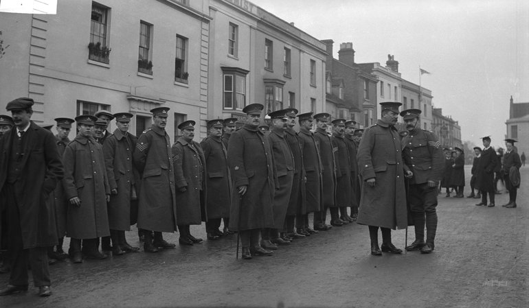 First World War soldiers drawn up in ranks in Bridge Street, with a sergeant speaking to an officer in front of them. All are wearing flat hats and greatcoats.