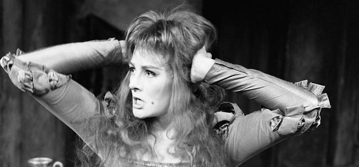 Katherine, her hair loose and wild and the laces of the bodice of her gown loosened, holds her hands to her head as she snarls (presumably at Petruchio).