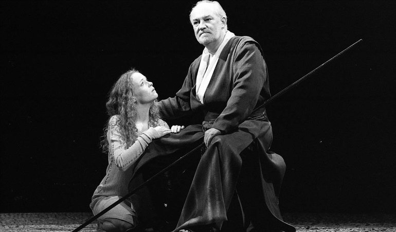 The Tempest Royal Shakespeare Company, 1998. A solemn-faced, white haired, seated Prospero stares out over the audience. On his right Miranda kneels, her hands on his thigh, staring up at him in concern. She has long curly hair and a light dress, contrasting with his dark robe with a long white collar.