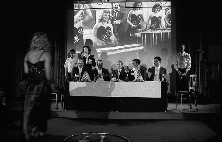 Timon's  banquet for Alcibiades. Four men and a woman in dark suits sit behind a table covered in a white cloth, and with rich ornaments on it. Servants stand behind them. In front, with her back to the audience, is a woman in a skimpy black dress who is clearly part of the entertainment; four other similarly-dressed women, playing two large xylophones, are projected on a screen behind the diners.