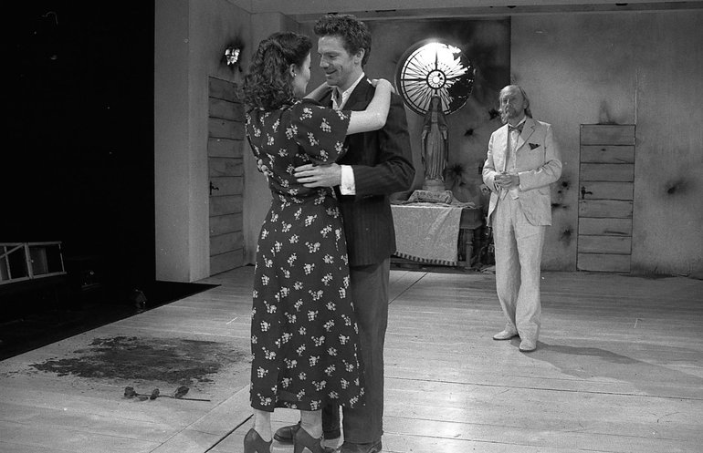 In Edwardian dress, a dark-suited Troilus holds the waist of Cressida, in a dark dress with a white design, whose arms are on Troilus's shoulders. A white-suited Pandarus stands in the background looking on.