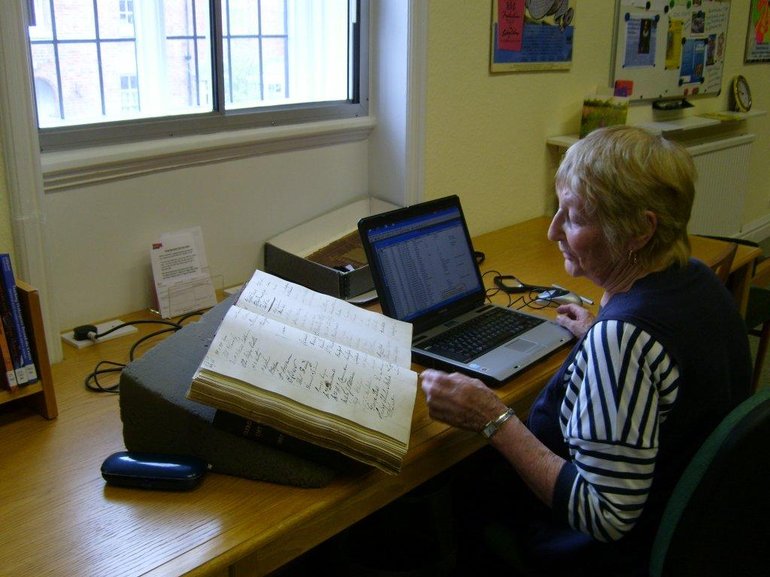 A woman sits at a desk in front of a laptop making notes. To her left is an old book from the Shakespeare Birthplace Trust's collection which she is reading.