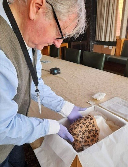 Volunteer Jon inspecting a woman’s cap from the 1600s. It is peach-coloured with black embroidered decoration. He is lifting out of a box lined with soft tissue, wearing purple latex gloves.