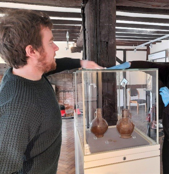 Volunteer Will assisting with an exhibition installation, cleaning a glass display case inside which are two small brown earthenware jugs.